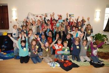 Ökumenische Flugreise für Kinder ins Weltgebetstagsland Viele Kinder sitzen auf dem Boden eines Pfarrsaals, umgeben von bunten Tüchern. Alle strecken die Hände nach oben, ein Kind hält einen Globus in die Höhe